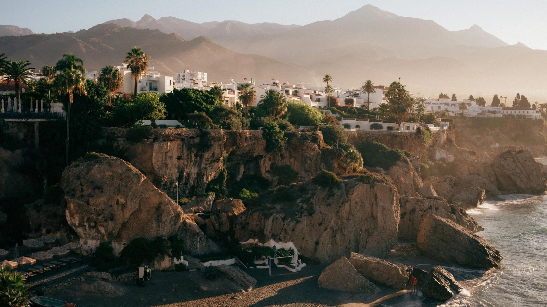 White houses and palm trees on the cliffs above a small beach at Nerja, with mountains behind in the early light