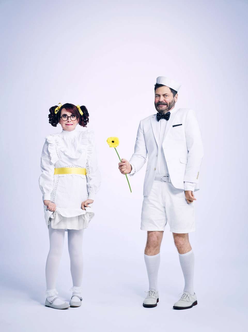 Megan & Nick dresses up as young children in all white clothing. Both looking awkward. Nick holding a yellow flower.