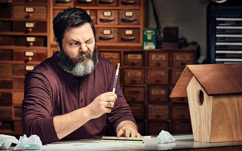 Nick Offerman with a pencil in hand looking at a wooden bird house.