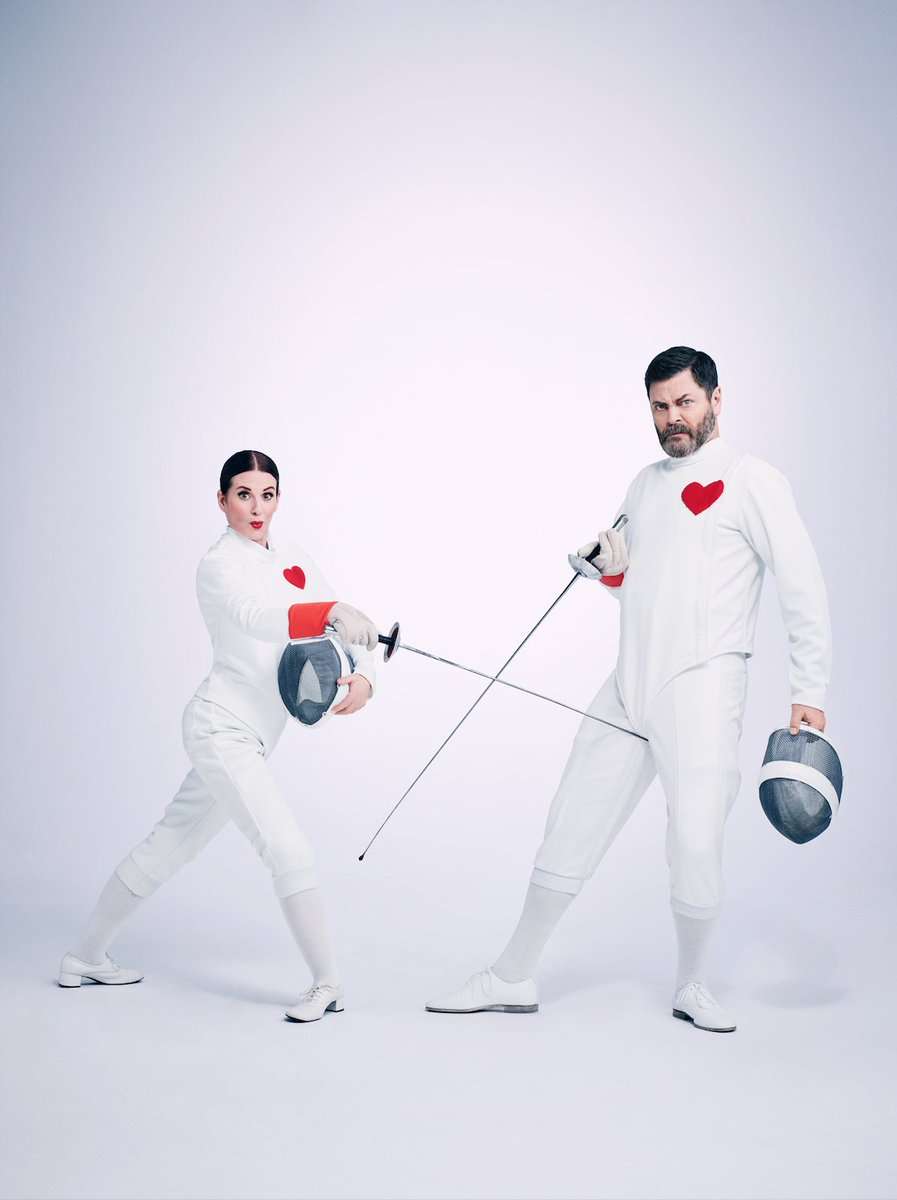 Megan Mullally & Nick Offerman in fencing attire with red heart badges. Megan is poking Nick in the groin with her sword.