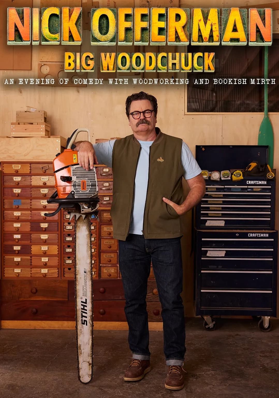Nick Offerman standing in front of storage furniture resting his arm on a huge chainsaw. He wears a brown vest over a light blue t-shirt. 