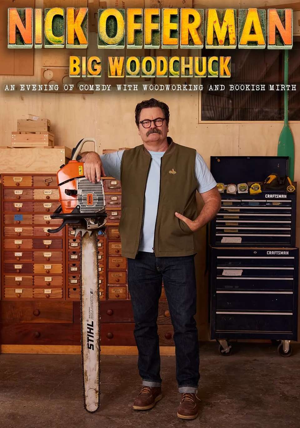 Nick Offerman standing in front of storage furniture resting his arm on a huge chainsaw. He wears a brown vest over a light blue t-shirt.