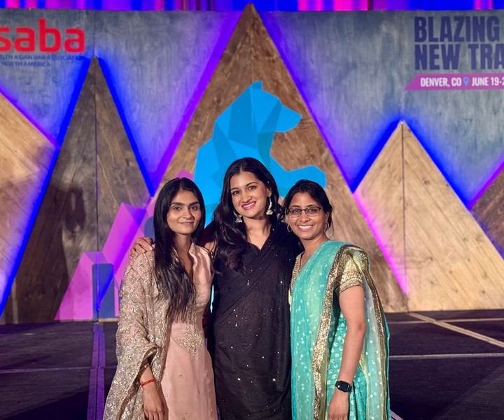 Three attendees in South Asian formal wear smile on stage in front of a ‘Blazing New Trails’ backdrop at SABA North America’s 2025 conference in Denver