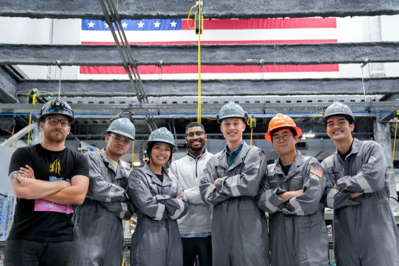 Gecko team in hard hats and coveralls beneath a U.S. flag in Gecko’s facility