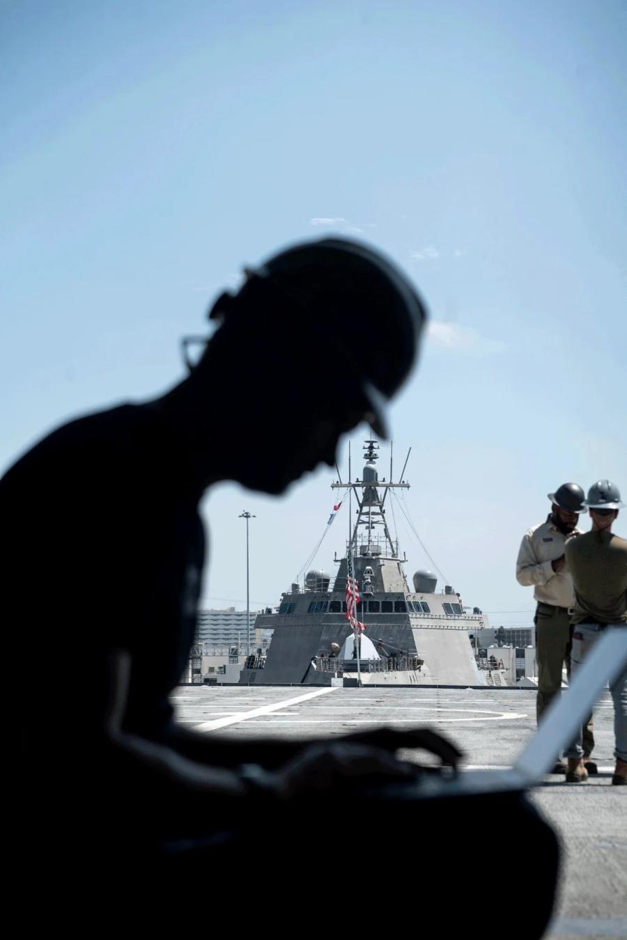 A Gecko engineer working on a laptop on a ship deck, a Navy ship behind