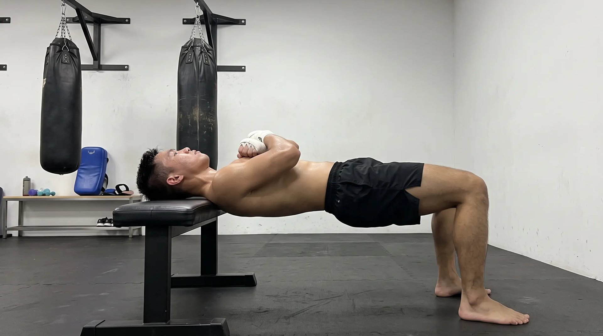 A fighter performs an isometric neck plank, bridging his body in a straight line with only the back of his head and shoulders supported on a gym bench, in a fight gym with heavy bags behind him.