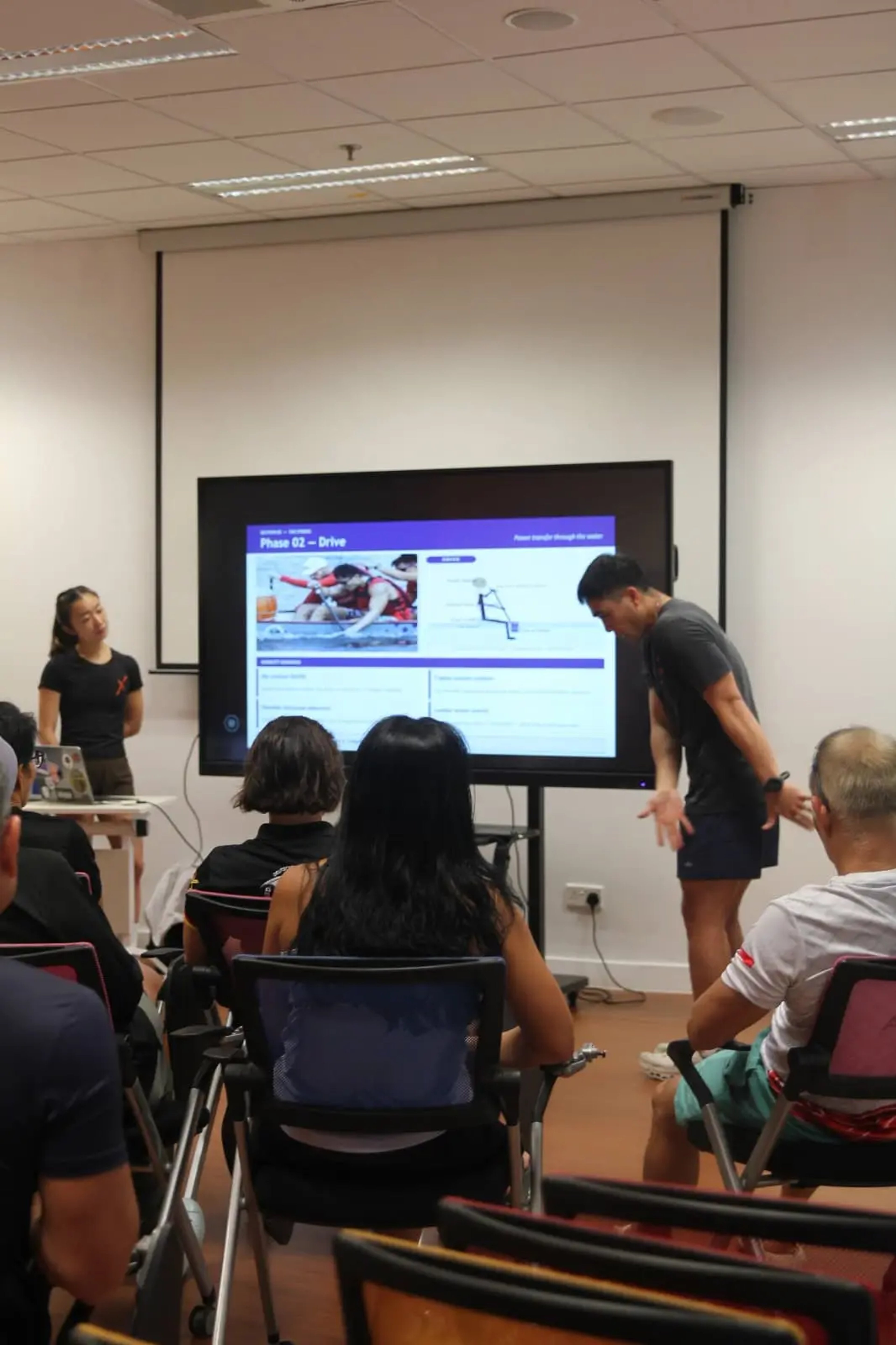 ActiveX Physio practitioners presenting a paddle sports workshop. Isabelle stands beside a laptop on the left, while Nicholas Ho presenting in dark athletic wear demonstrates a movement technique to the Singapore National Dragon Boat Senior Team. The screen behind them displays "Phase 02 — Drive" with dragon boat imagery and coaching cues on force transfer through the water.
