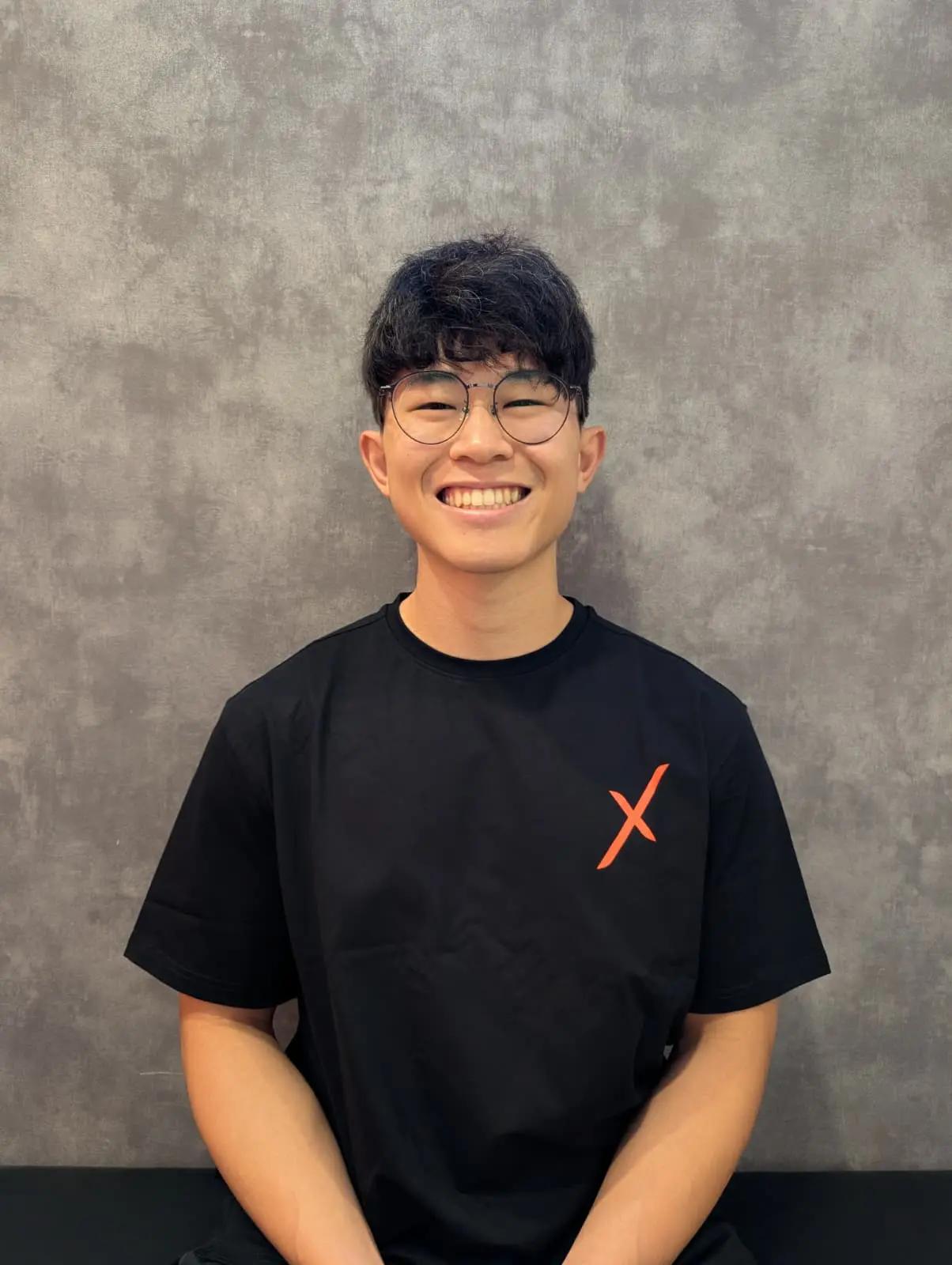 Daniel Ng smiling in a black Activex Physiotherapy t-shirt with the orange X logo, standing against a grey textured wall.
