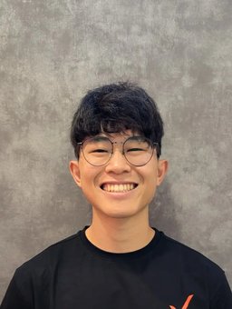 Daniel Ng smiling in a black Activex Physiotherapy t-shirt with the orange X logo, standing against a grey textured wall.