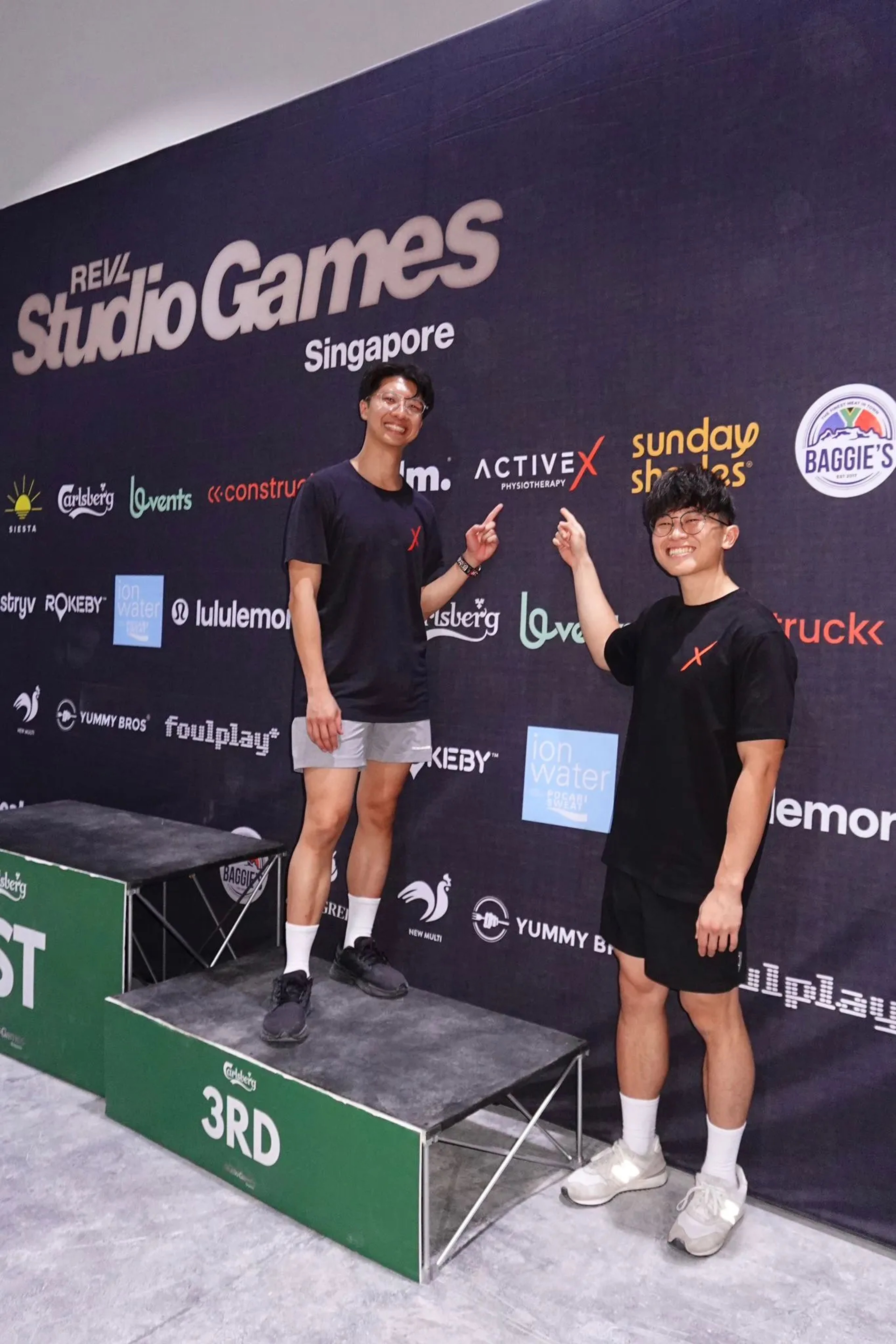 ActiveX Physio practitioners Ivan Tam and Daniel Ng at the REVL Studio Games Singapore, both in black ActiveX-branded t-shirts, grinning and pointing up at the ActiveX Physiotherapy logo on the sponsor backdrop behind them. One stands on the 3rd-place podium block. The backdrop features sponsors including lululemon, Carlsberg, Ion Water, and Foulplay.