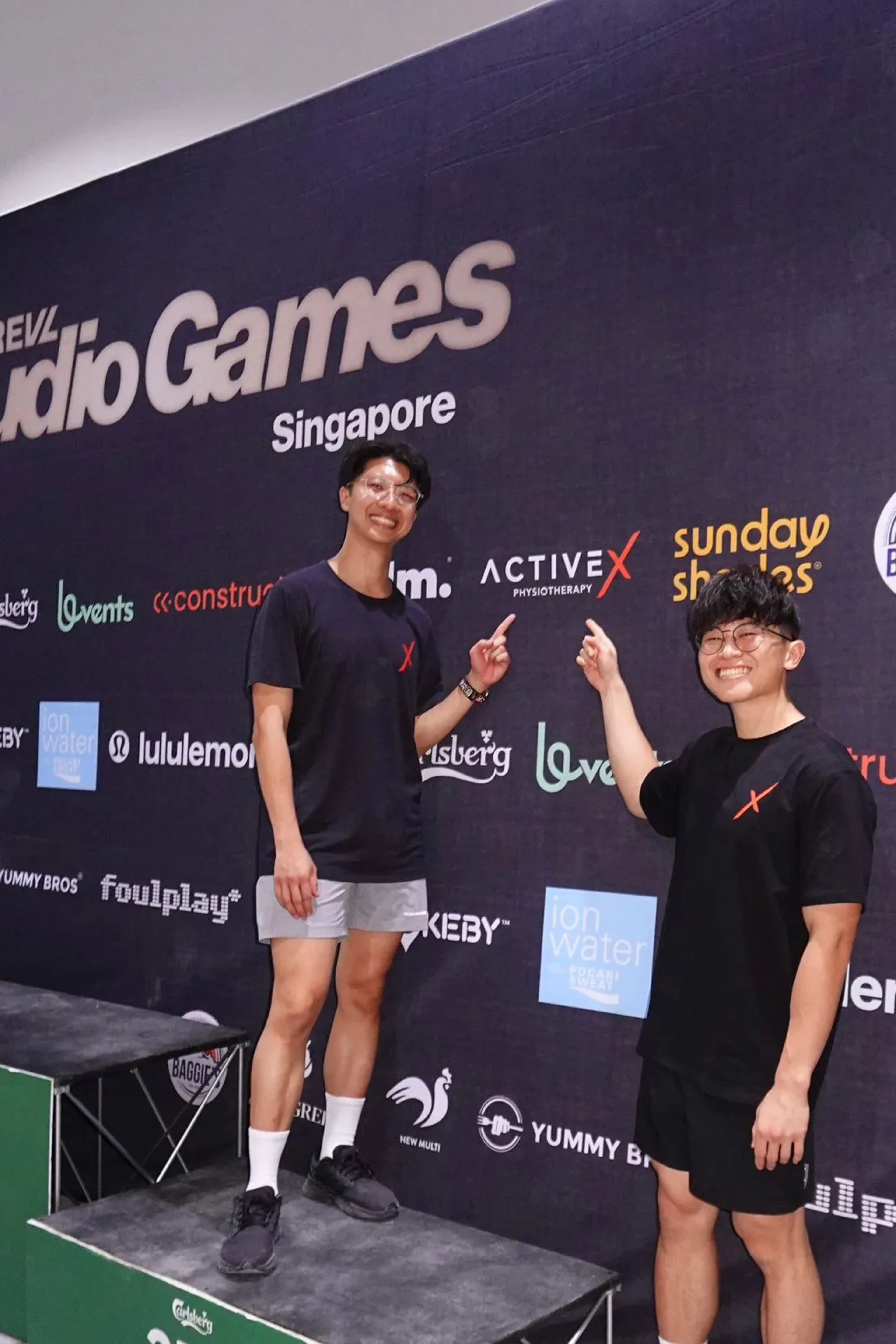 Activex Physio practitioners Ivan Tam and Daniel Ng at the REVL Studio Games Singapore, both in black Activex-branded t-shirts, grinning and pointing up at the Activex Physiotherapy logo on the sponsor backdrop behind them. One stands on the 3rd-place podium block. The backdrop features sponsors including lululemon, Carlsberg, Ion Water, and Foulplay.