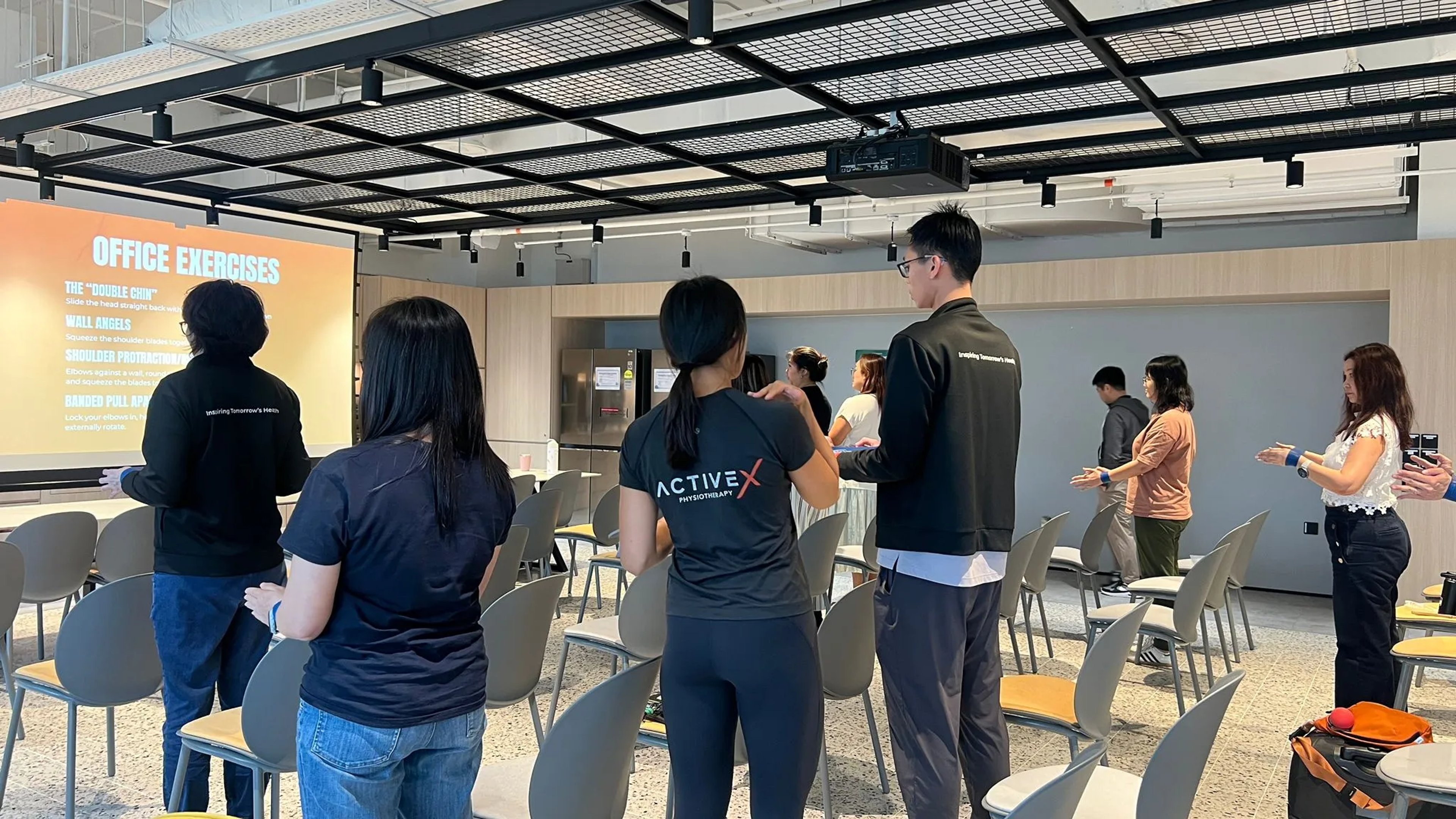 ActiveX Physiotherapy team leading a corporate office exercises workshop, with practitioners in branded black ActiveX shirts guiding a group of office workers through shoulder and posture exercises. A presentation slide titled "Office Exercises" is displayed on screen, listing movements including the "Double Chin," Wall Angels, Shoulder Protraction, and Banded Pull Aparts.