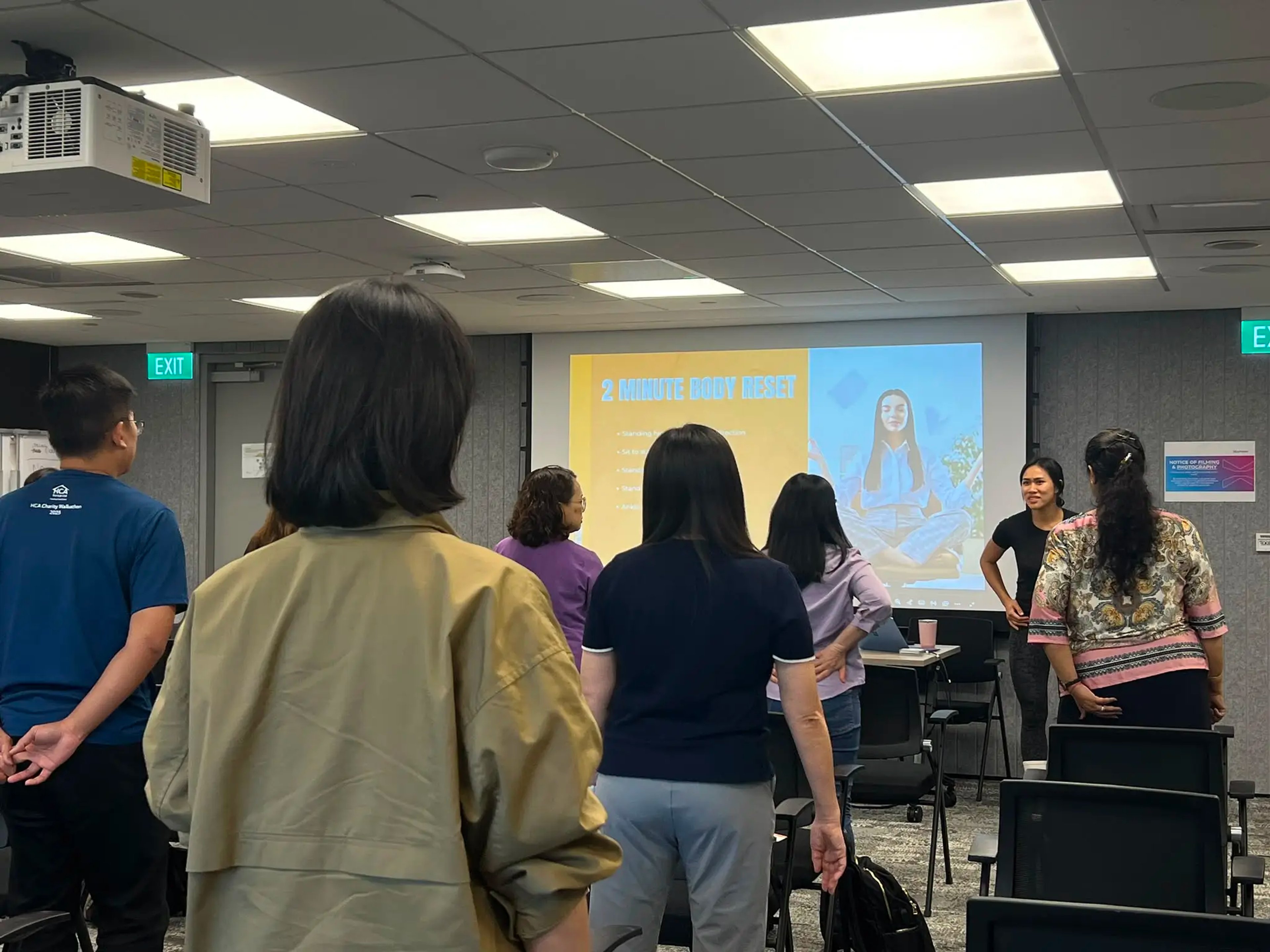 ActiveX Physio founder Helen Nguyen leading a standing movement exercise during a corporate wellness talk at Synapxe. Attendees are on their feet following along as the projector displays a "2 Minute Body Reset" slide. The session focused on preventative care for hip and knee pain.