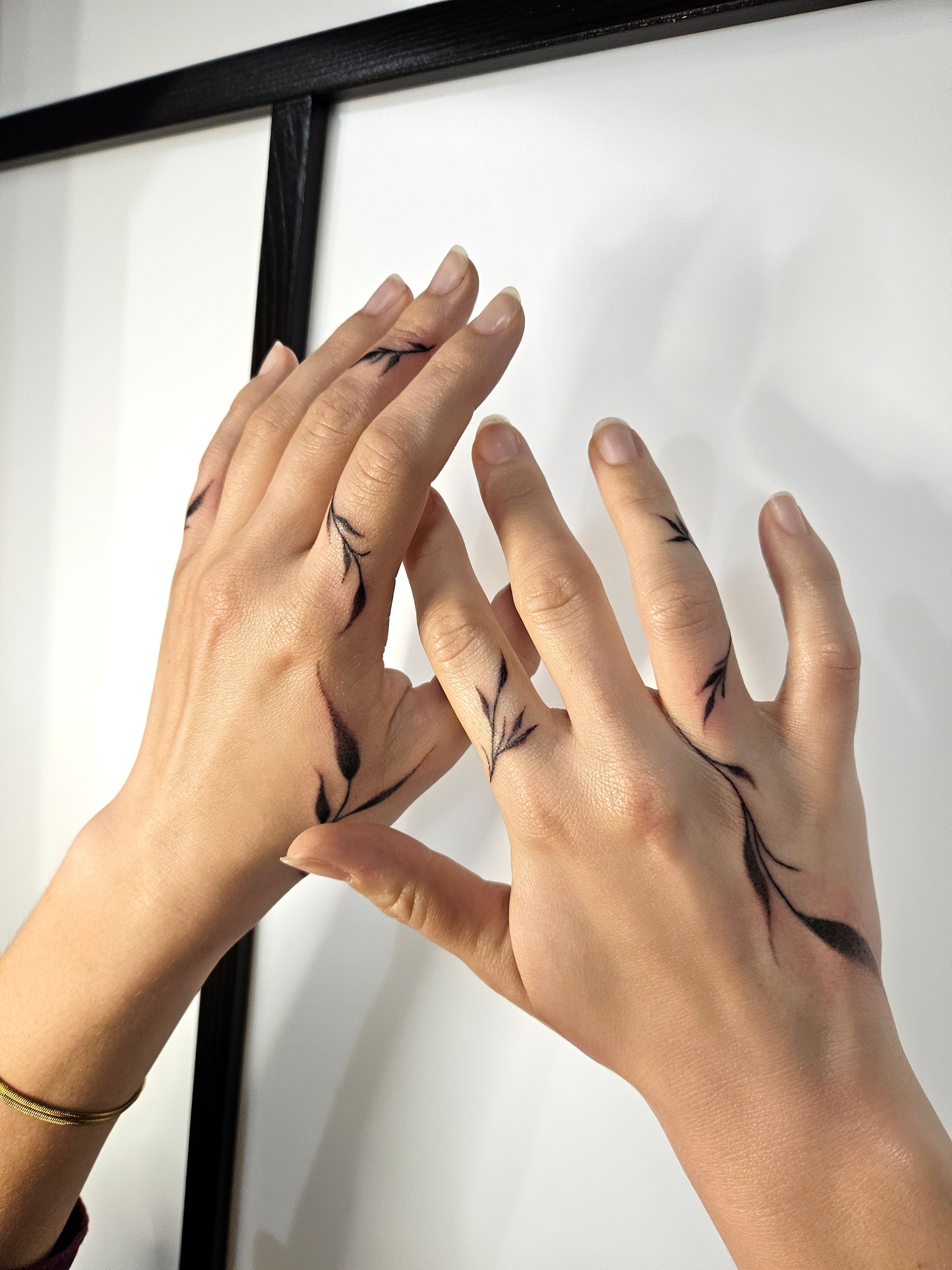 Tatouages végétaux noirs composés de fines branches et de feuilles effilées sur le dos des deux mains et les doigts.