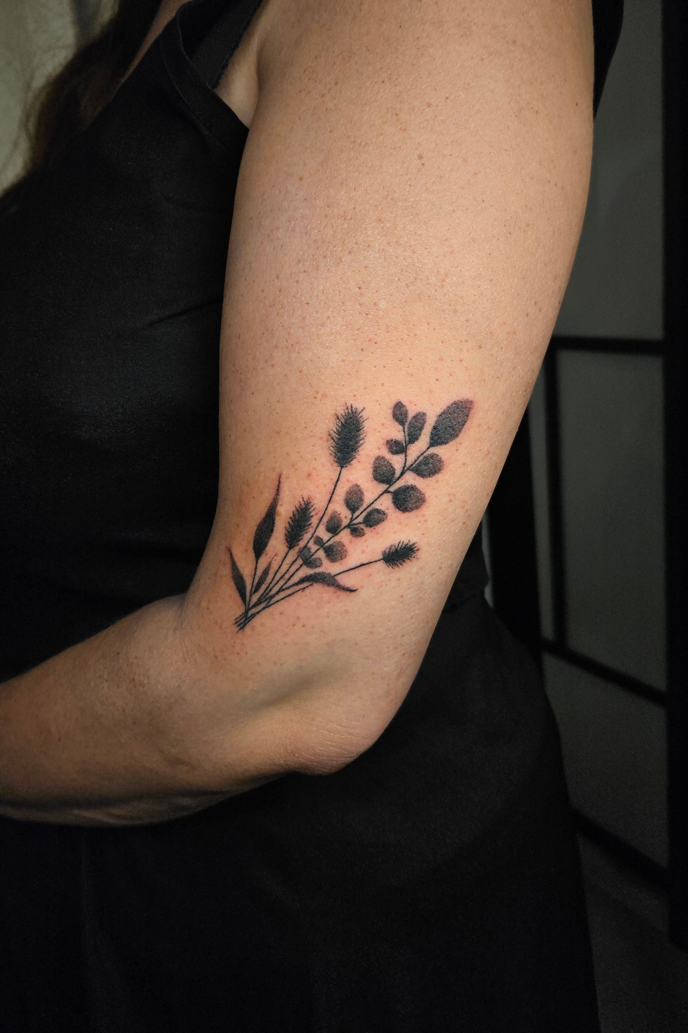 Tatouage botanique noir représentant un bouquet de tiges fines, de feuilles et d’épis sur le bras, près du coude.