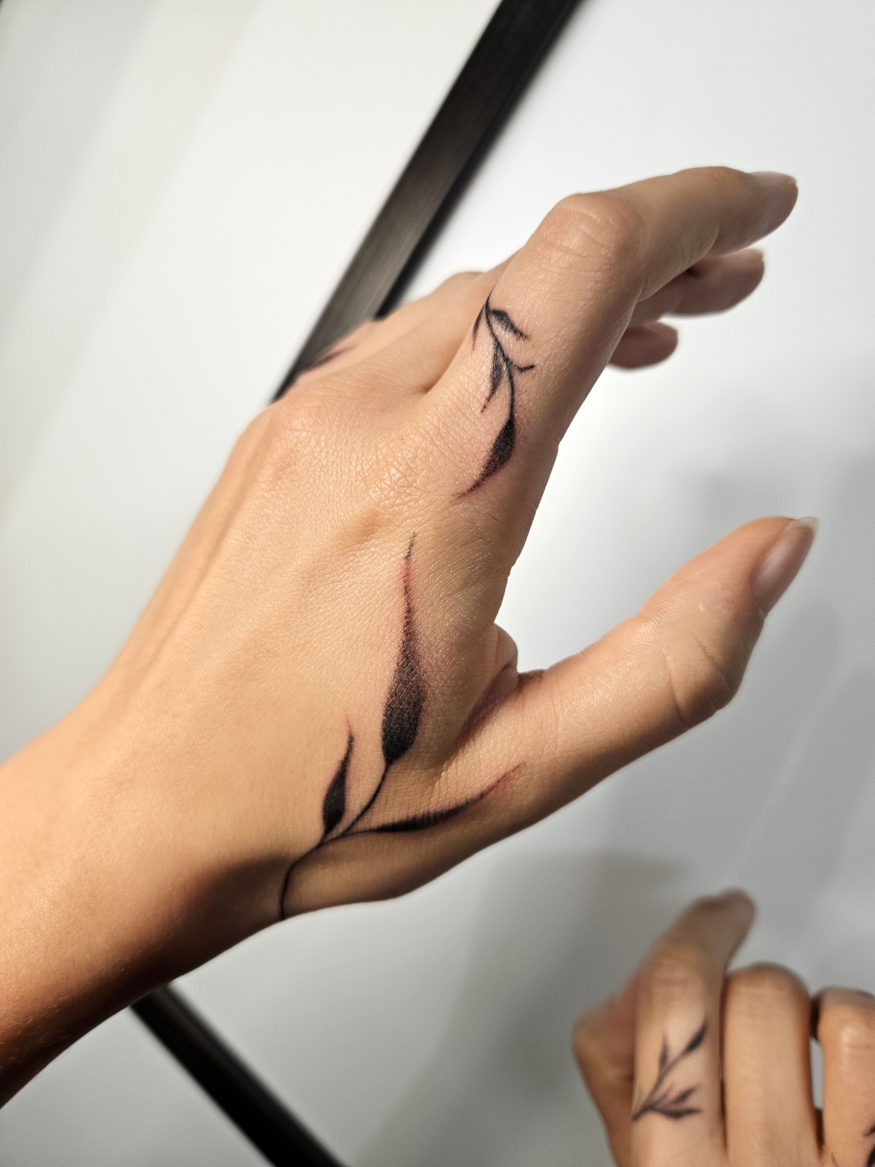 Vue rapprochée d’un tatouage végétal noir aux tiges courbes et aux feuilles effilées sur la main et l’index.