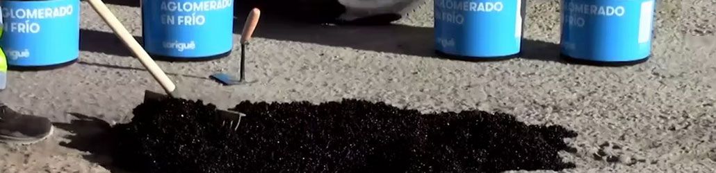 Toepassing van koudasfalt op een wegdek, met gereedschap en blauwe emmers met opschrift “Aglomerado en frío” zichtbaar op de achtergrond.