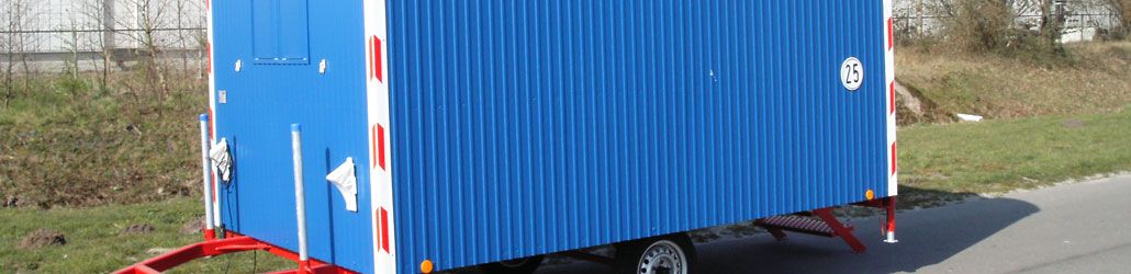 Blauwe bouwkeet op wielen met rode onderstel, geparkeerd langs de weg bij een grasveld.