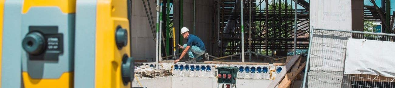 Bouwvakker met veiligheidshelm werkt op een bouwplaats en gebruikt een meetinstrument, met op de voorgrond een geel bouwlaser-apparaat.