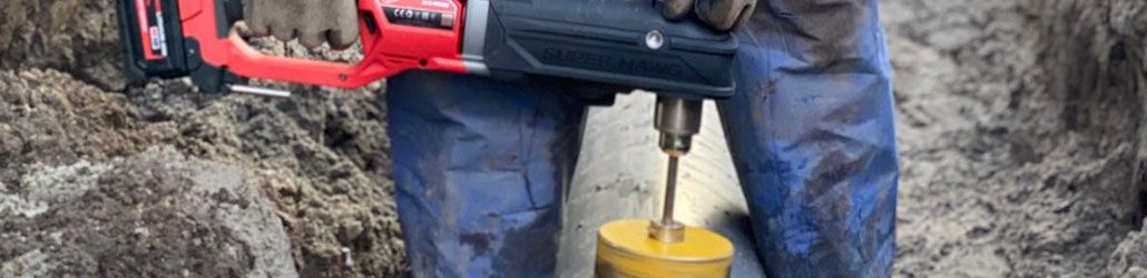 Monteur boort met een Milwaukee gatenzaag een opening in een kunststof leidingbuis op een modderige bouwplaats.