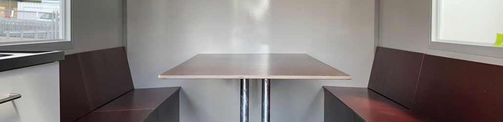 Interieur van een schaftkeet met twee houten banken en een tafel tussenin, omgeven door witte wanden en ramen aan beide zijden.