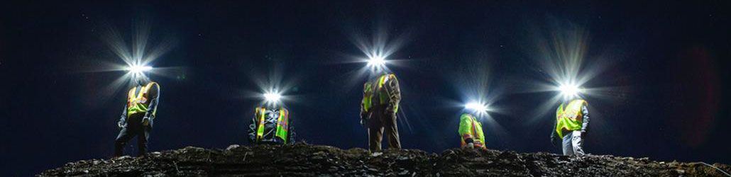 Zes bouwvakkers staan 's nachts op een verhoogd terrein, elk met een felle helmlamp en reflecterend veiligheidsvest. De helmlampen lichten krachtig op tegen de donkere hemel.