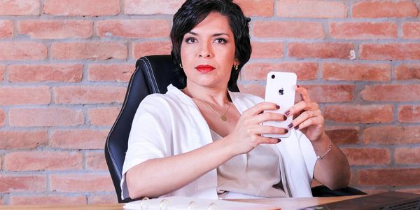 Professional woman using smartphone at desk with brick wall backdrop, representing remote work.