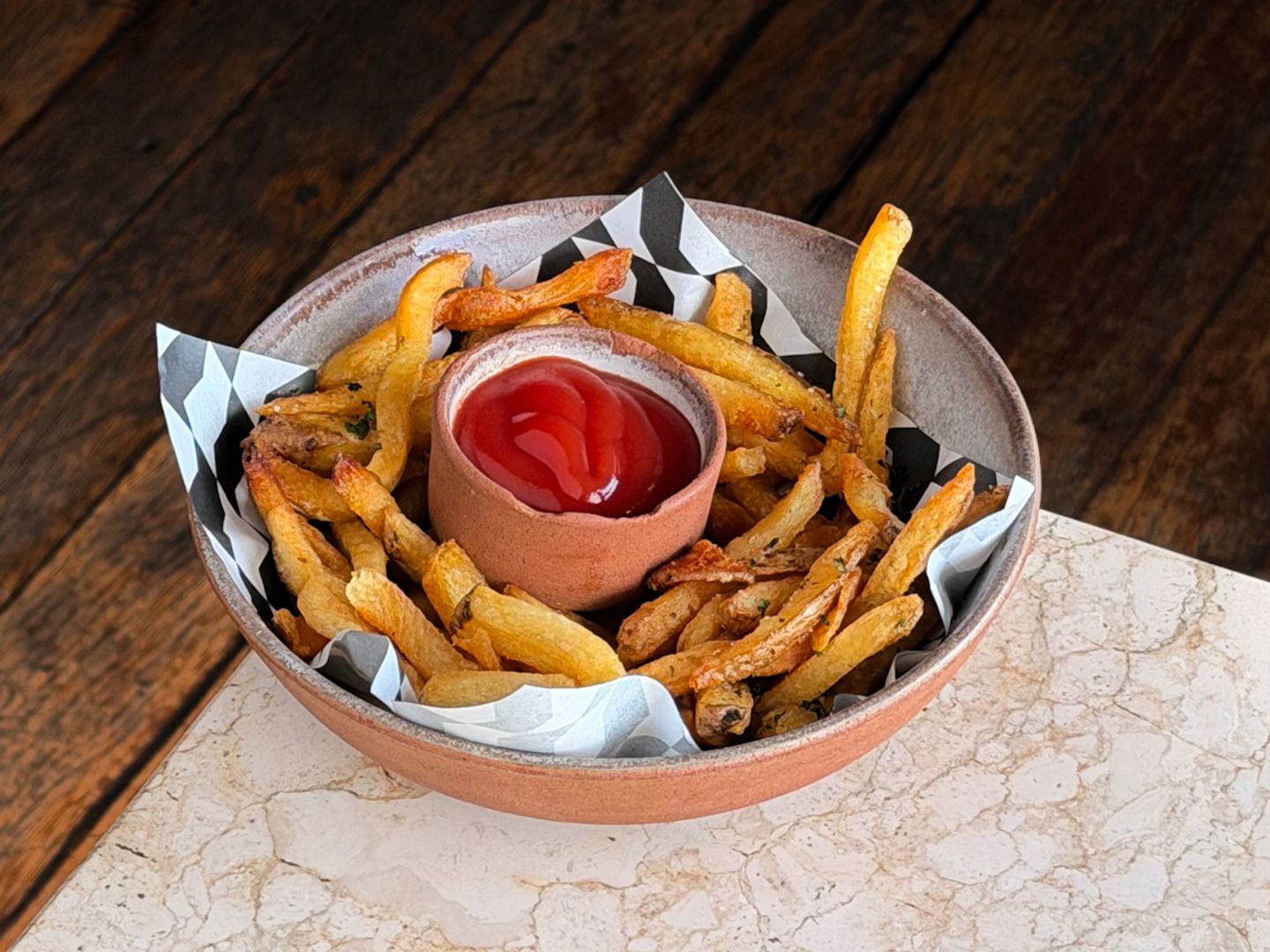 Crispy golden fries in a paper-lined ceramic bowl with a clay pot of ketchup in the centre.