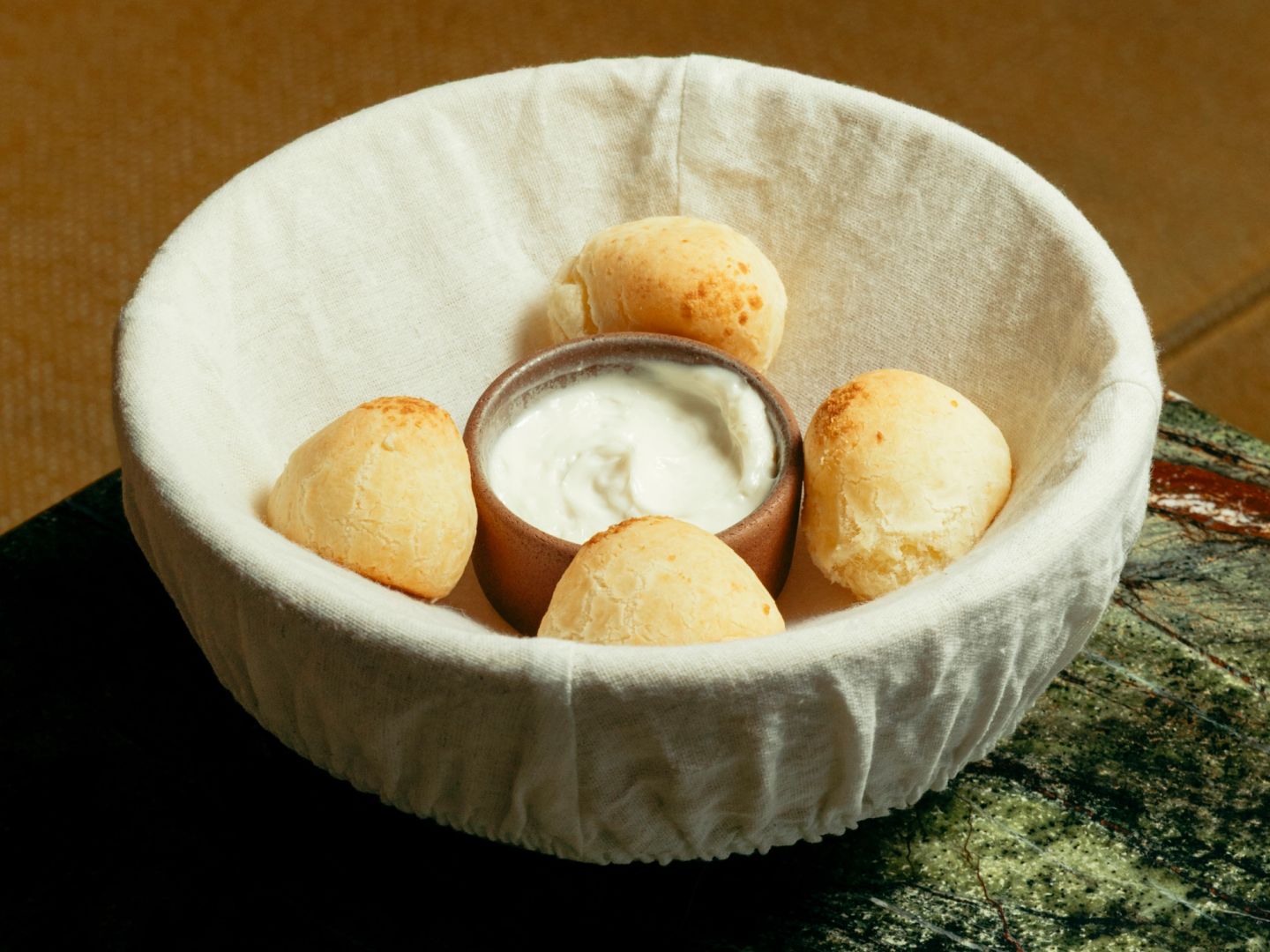 Golden Brazilian-style cheese bread balls in a linen-lined bowl around a clay pot of white dip.