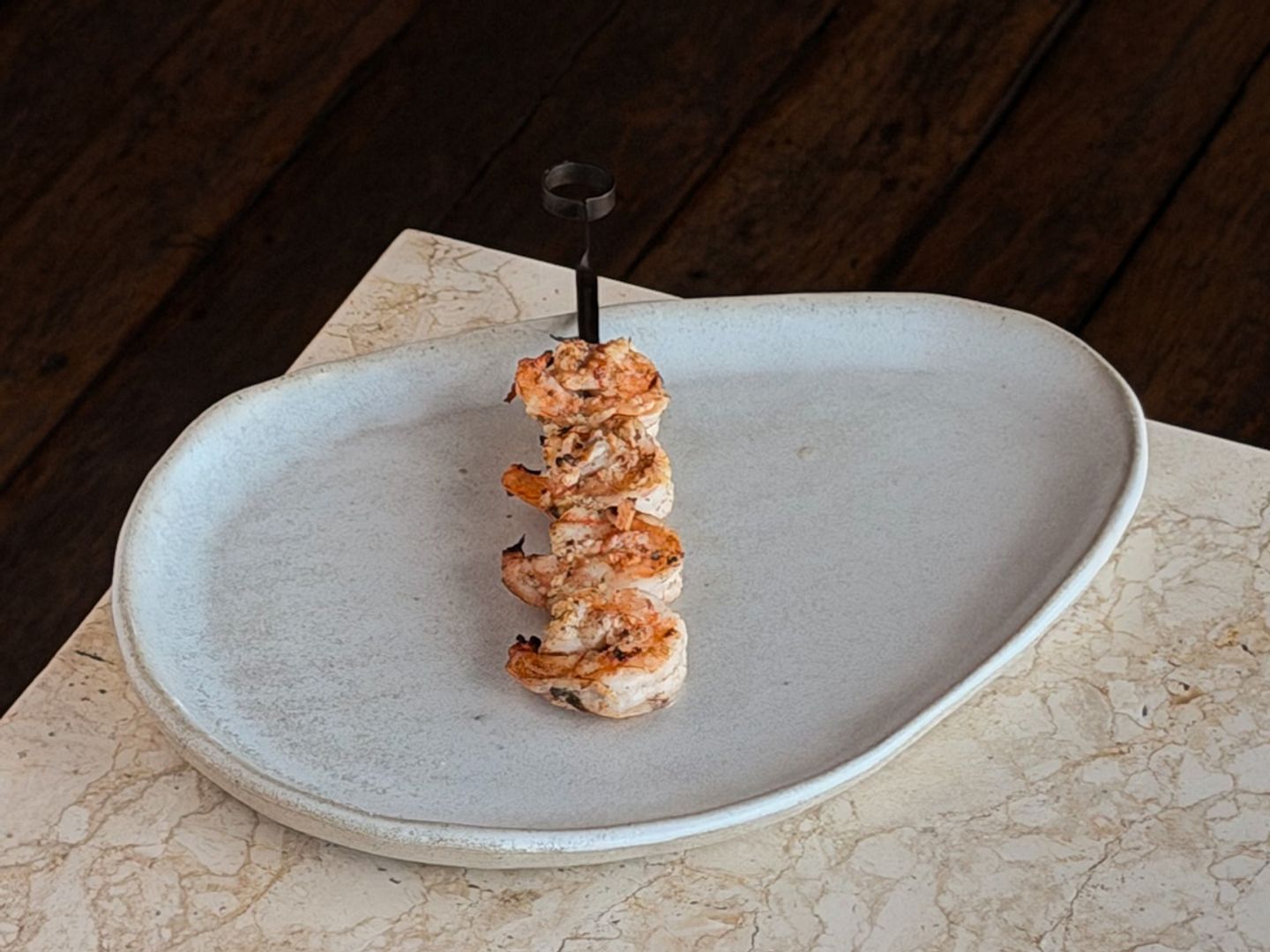 Grilled prawns threaded on a metal skewer standing upright on an oval stoneware plate.