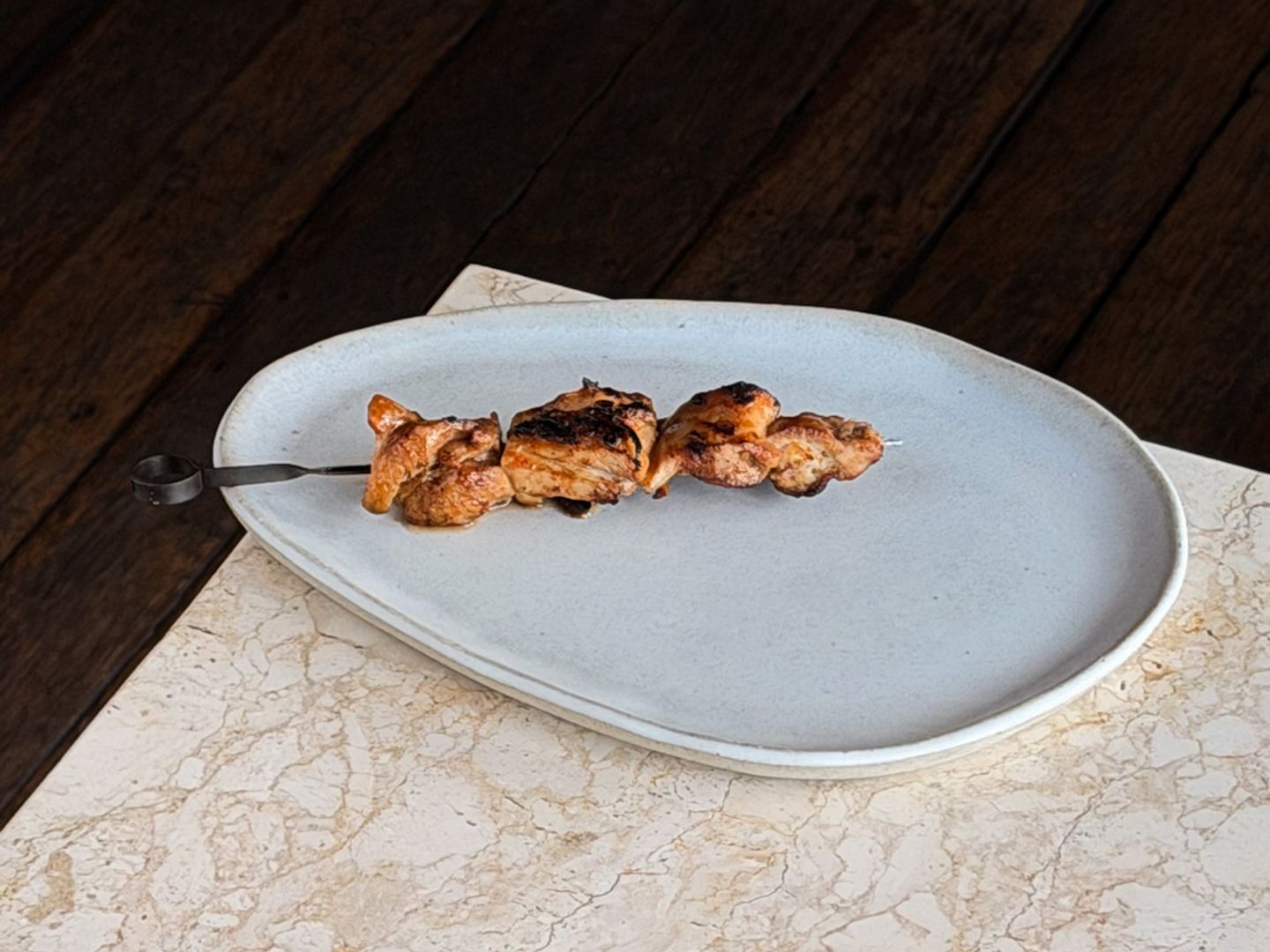 Grilled, charred chicken thigh pieces on a metal skewer set on an oval ceramic plate.