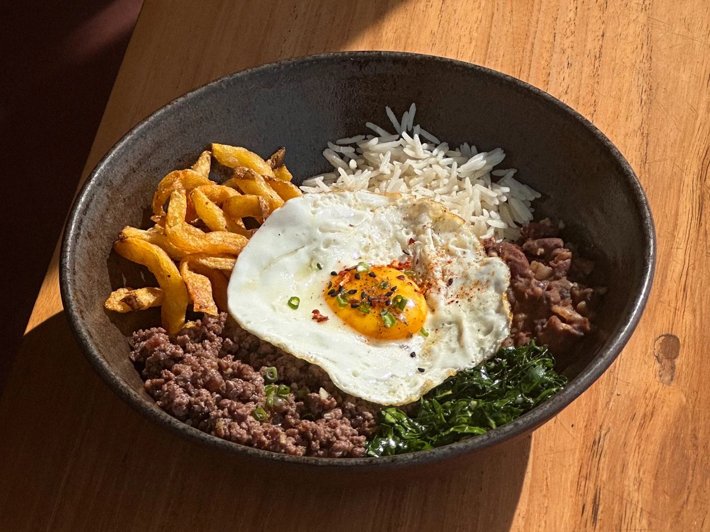  Ground beef and rice bowl with a fried egg, fries, and greens.