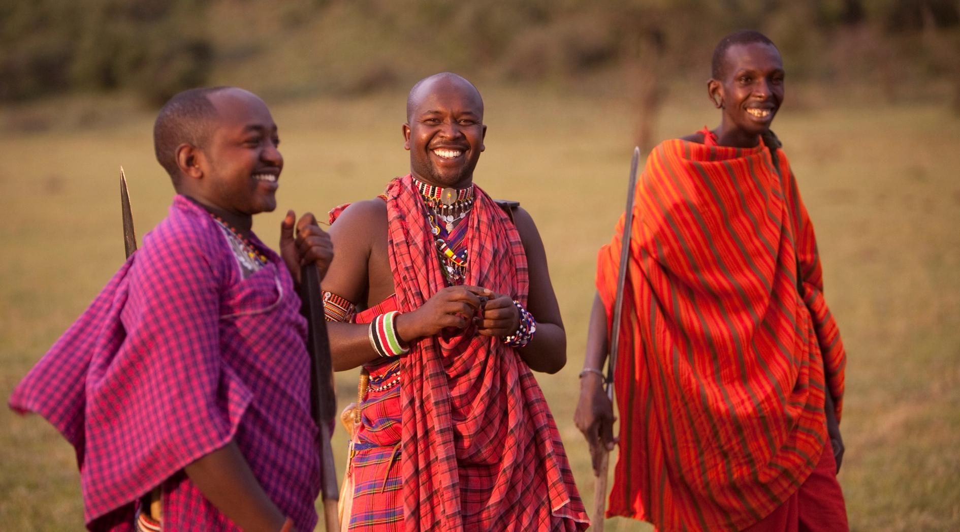 Three smiling Maasai men in traditional shukas (magenta, red, orange) holding staffs in a grassy field.