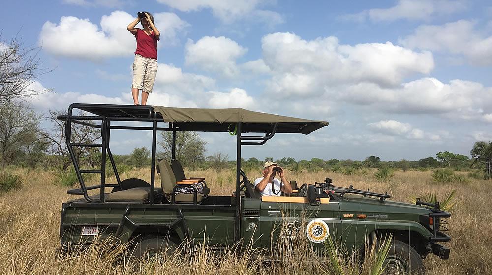 A woman stands on the roof of a safari vehicle and a man sits inside, both looking through binoculars in a grassy savanna.