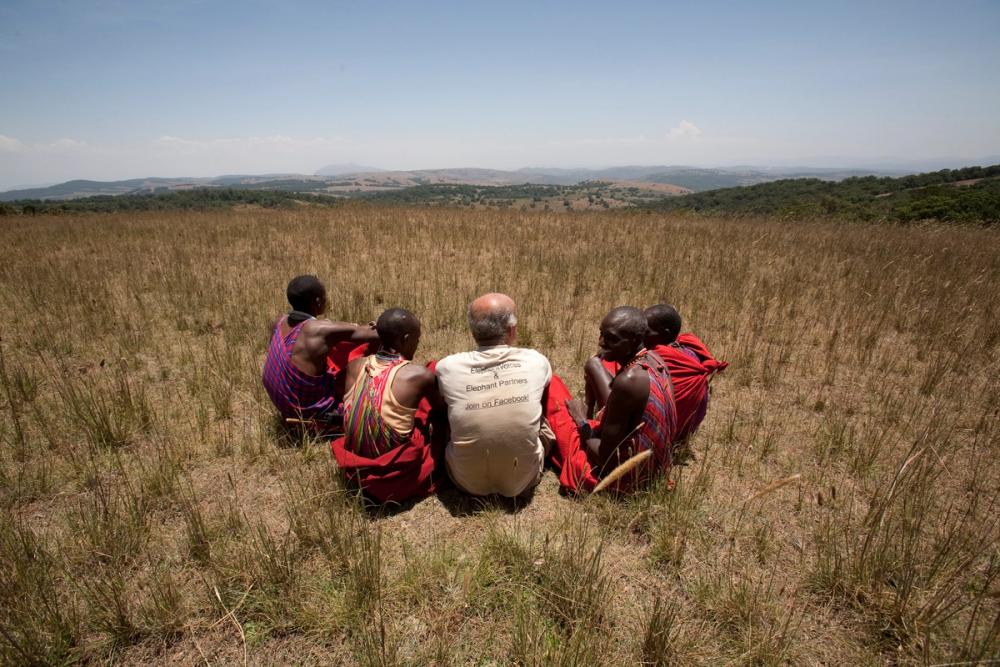 Six people, five in traditional red attire and one in a t-shirt, sit in a circle in a dry grassy field.