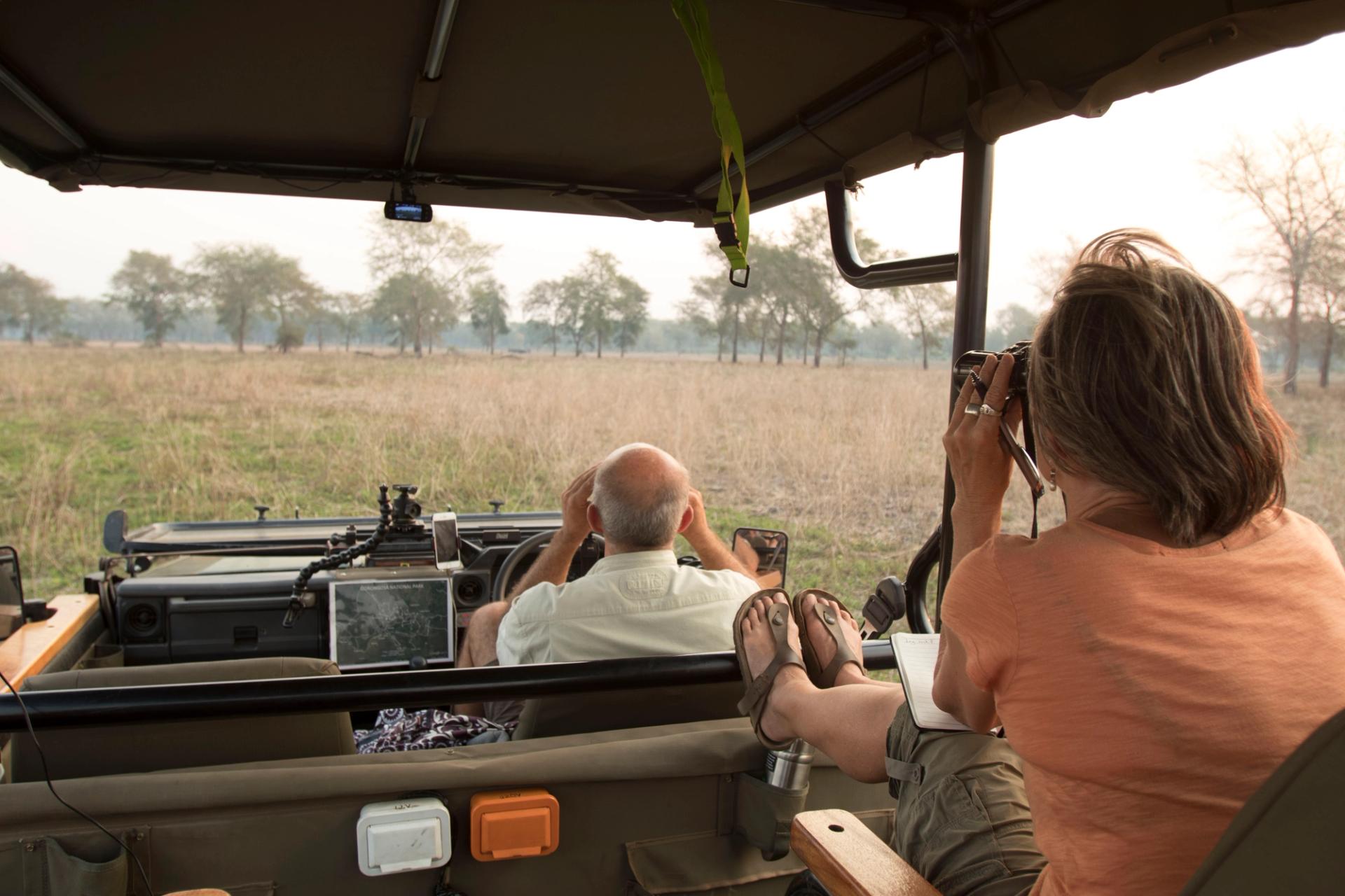 Two tourists in an open-top safari vehicle, one taking a photo, the other looking ahead.