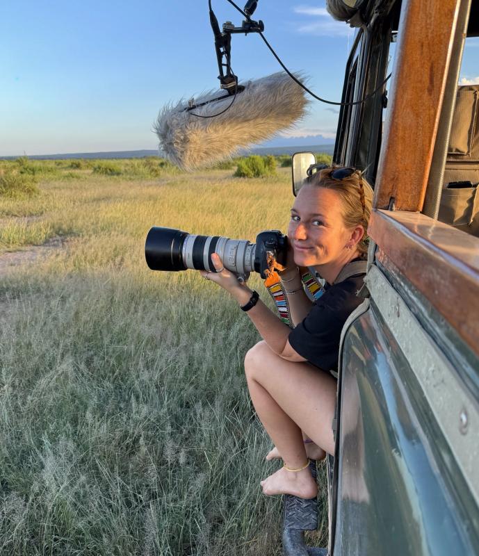 Selengei Poole-Granli leans out of a research vehicle holding a telephoto camera, with a fuzzy boom mic overhead, in a grassy savanna.