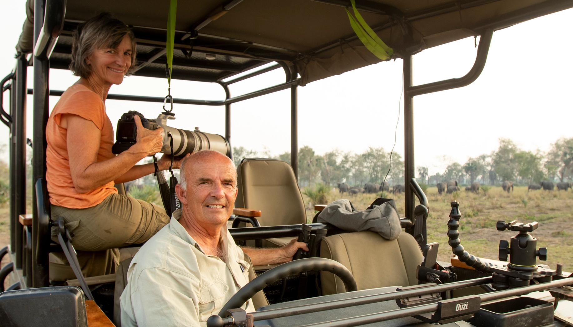 Smiling man and woman on a safari, the woman holding a telephoto camera, with a herd of animals in the background.