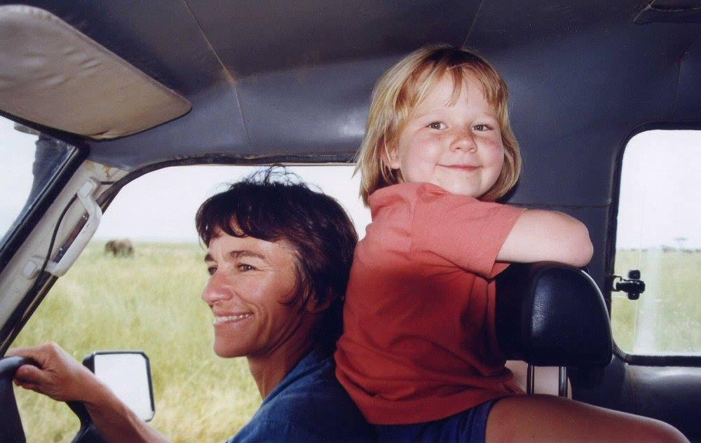 A woman drives a safari vehicle, smiling, while a child in a red shirt smiles at the camera, with an elephant in the grassy background.