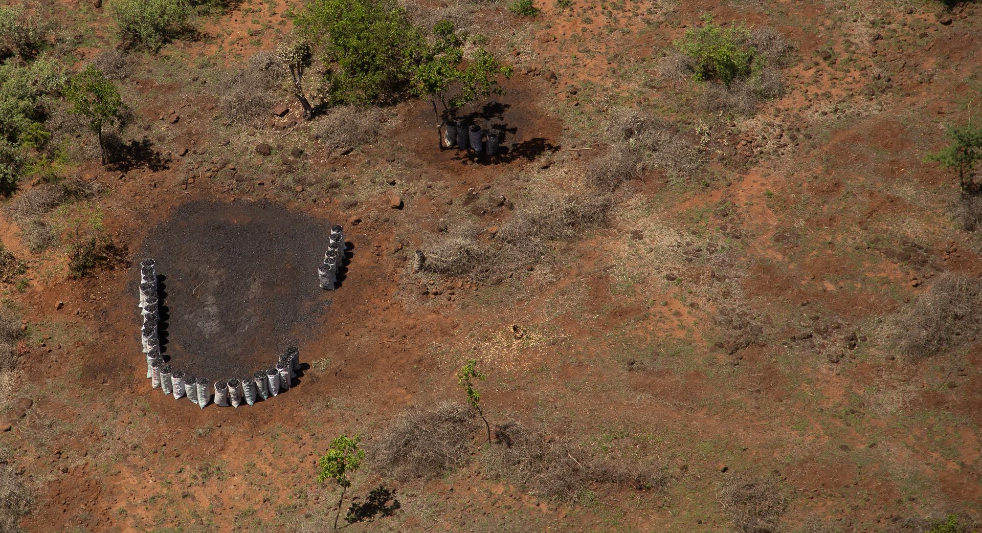 Aerial view of a dry, reddish landscape with a U-shaped arrangement of silver barrels surrounding a dark circular ground patch, and a small pile of barrels under a distant tree.