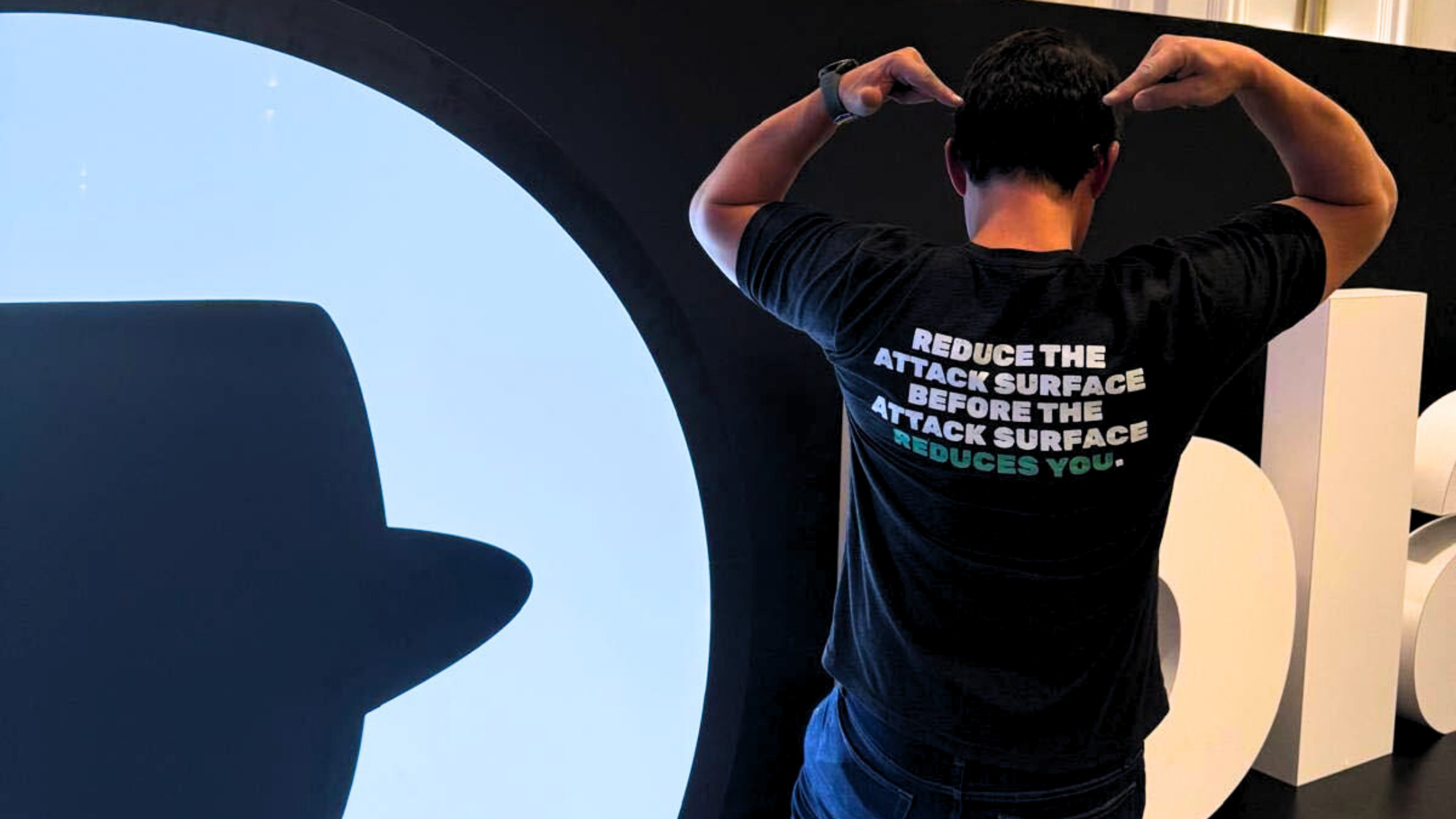 MagicSword team member posing in front of the Black Hat logo wearing a dark t-shirt that reads "Reduce the attack surface before the attack surface reduces you" - photographed from behind at Black Hat 2026.