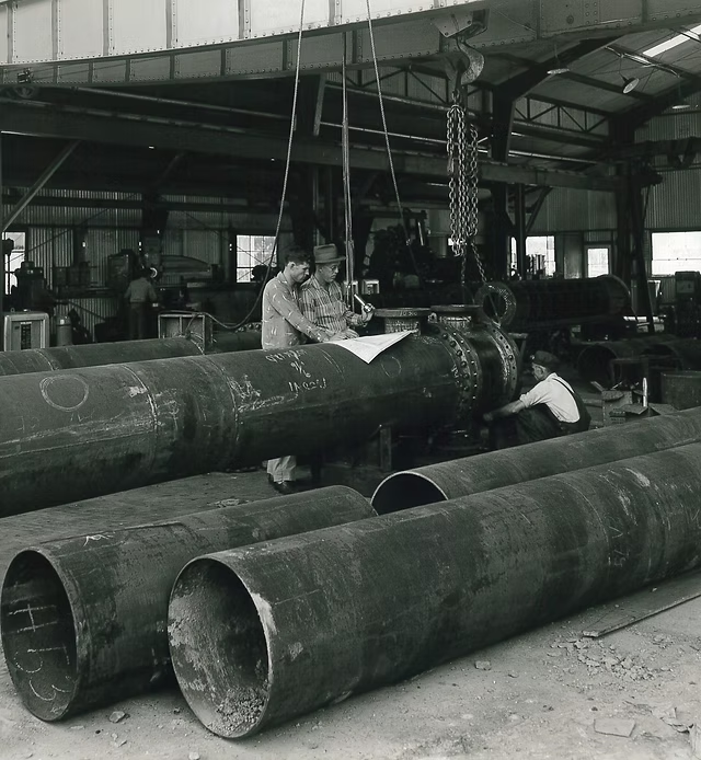 historical scene of industrial manufacturing, specifically the fabrication of large steel pipes inside a factory, likely during the mid-20th