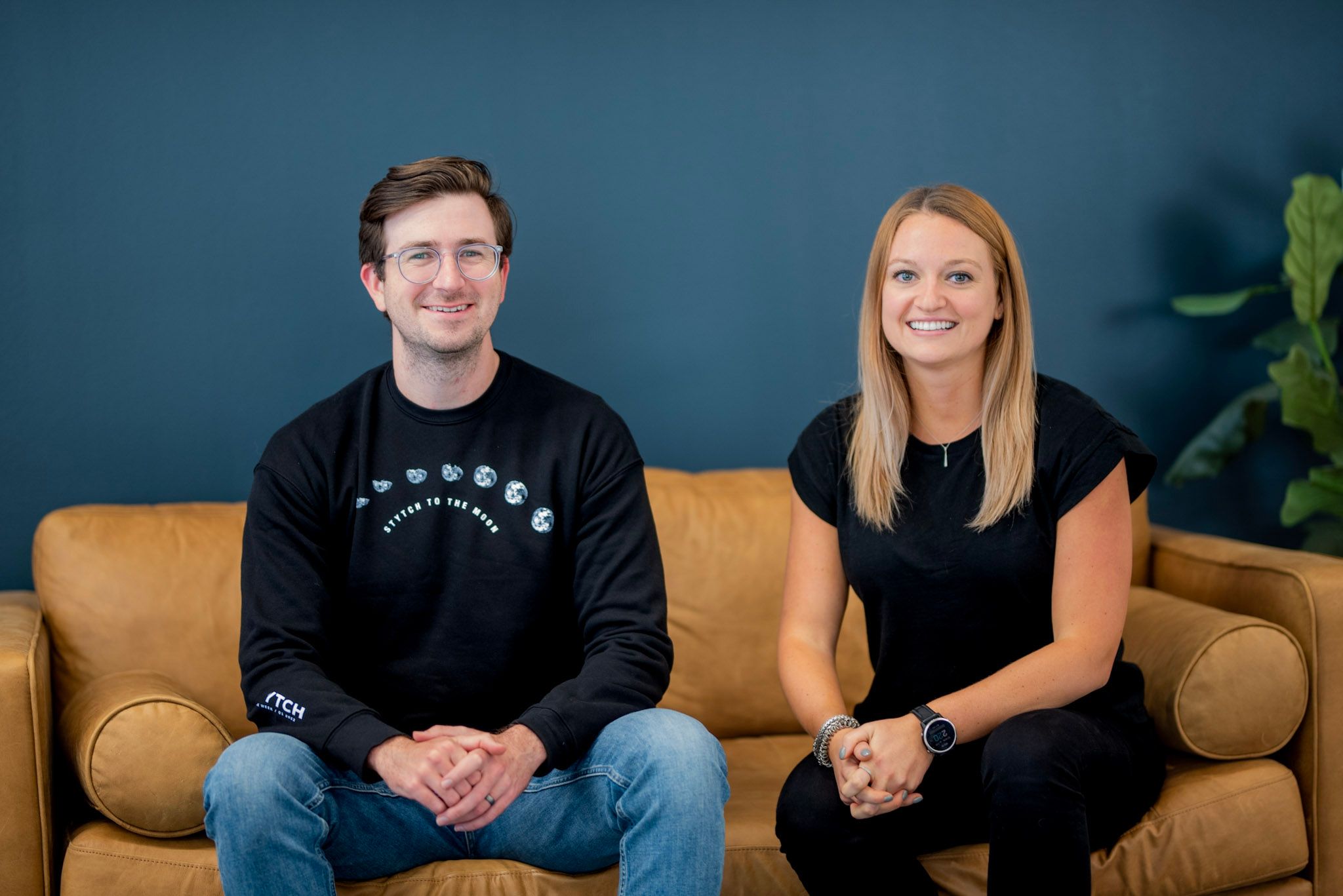 Photo of Stytch founders Reed McGinley-Stempel and Julianna Lamb, seated on a tan leather couch, both in black tees.