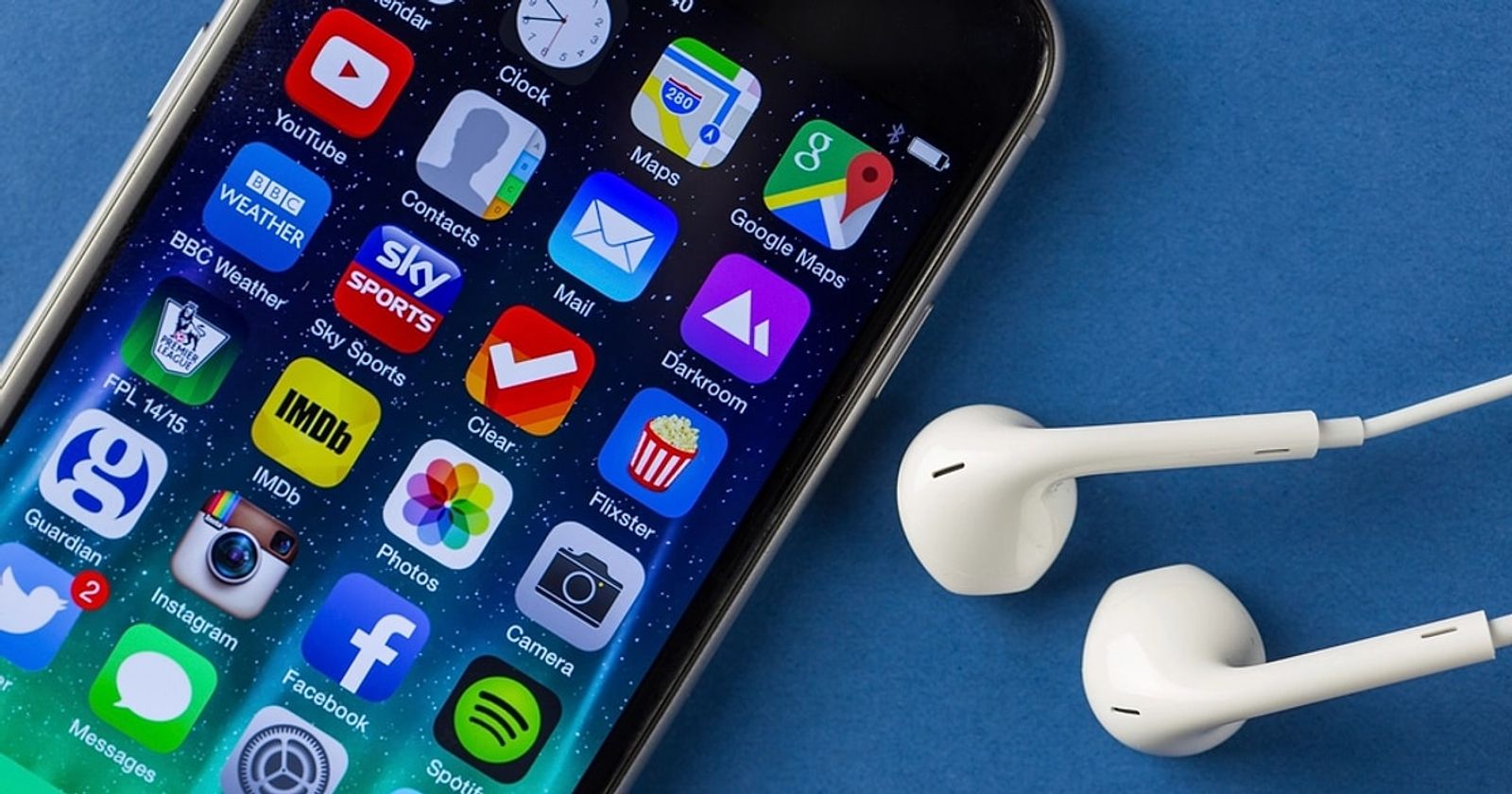 An iPhone home screen filled with app icons, lying at an angle beside a pair of white EarPods on a blue surface