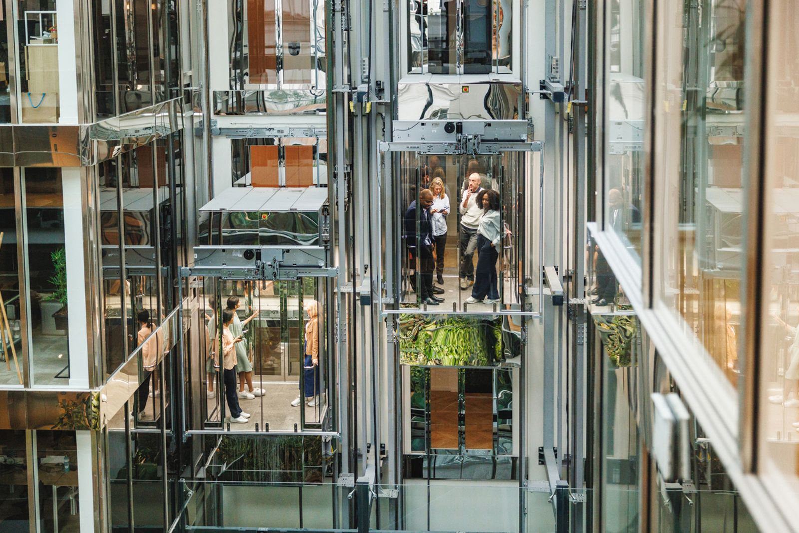 Men and women in professional attire engaging in discussion within the transparent lifts of a bustling, modern office environment.