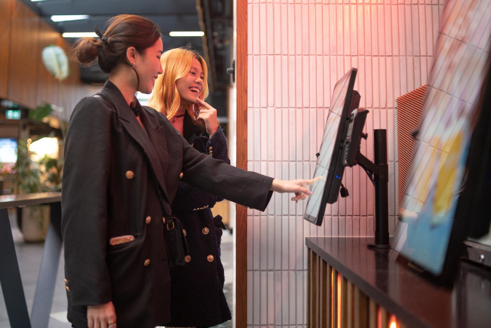 Two women order food and drinks on a touch screen kiosk