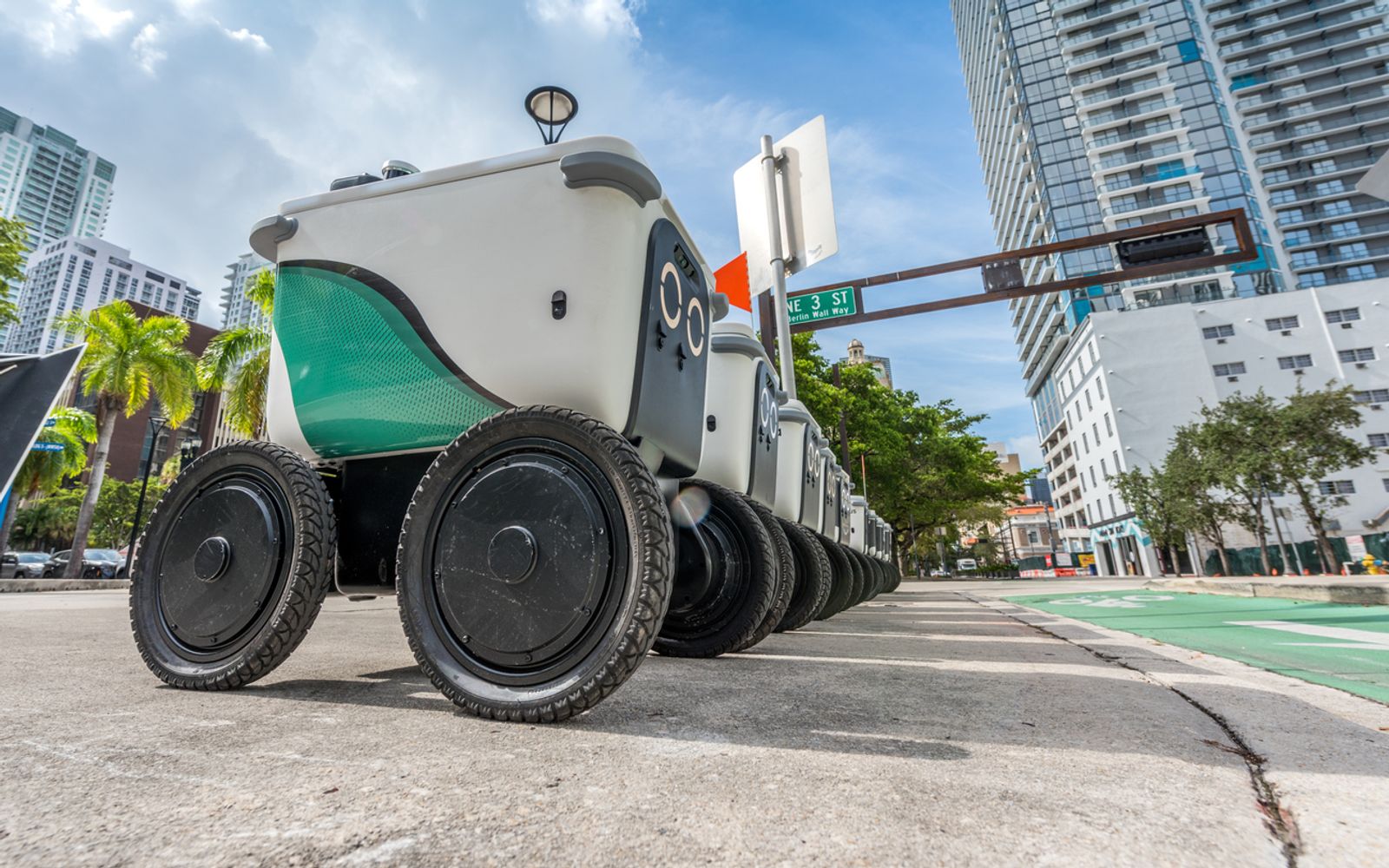 A fleet of delivery robots on a street corner in Downtown Miami awaits remote orders.