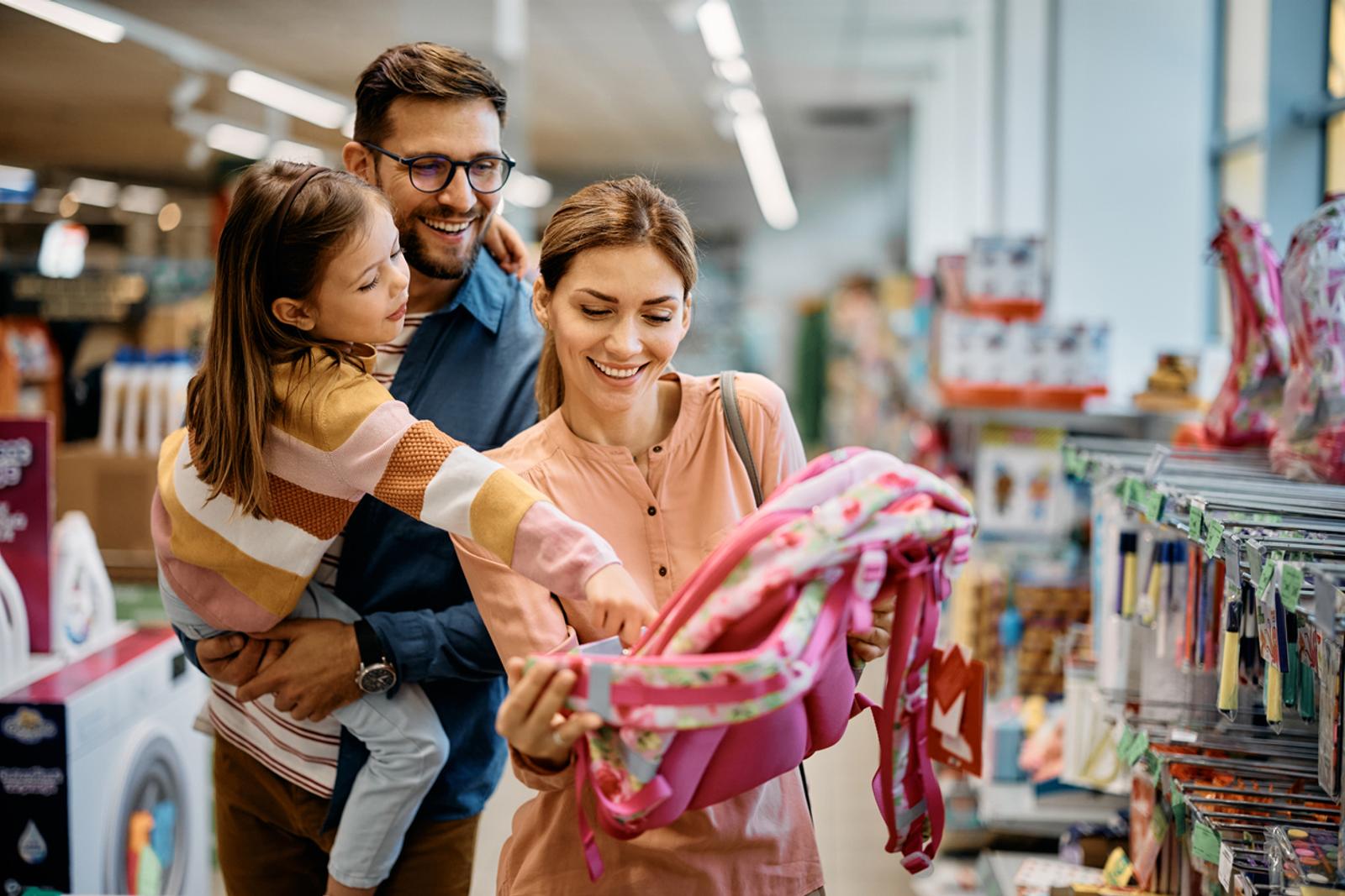 Young girl pointing at backpack while buying school supplies with her parents in supermarket