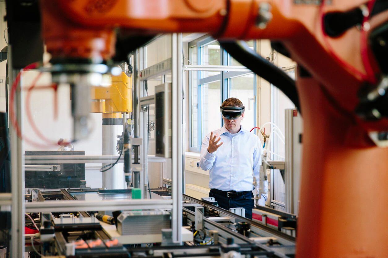 A man wears a VR helmet and uses his hand to control a robotic arm in a smart factory.