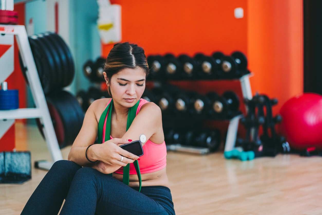 A young woman scans her glucose meter with a smart phone while sitting on the floor in a gym